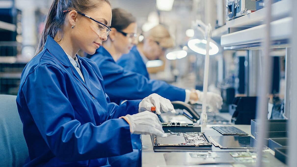 Female Electronics Factory Worker in Blue Work Coat and Protective Glasses is Assembling Laptop's Motherboard with a Screwdriver. High Tech Factory Facility with Multiple Employees.